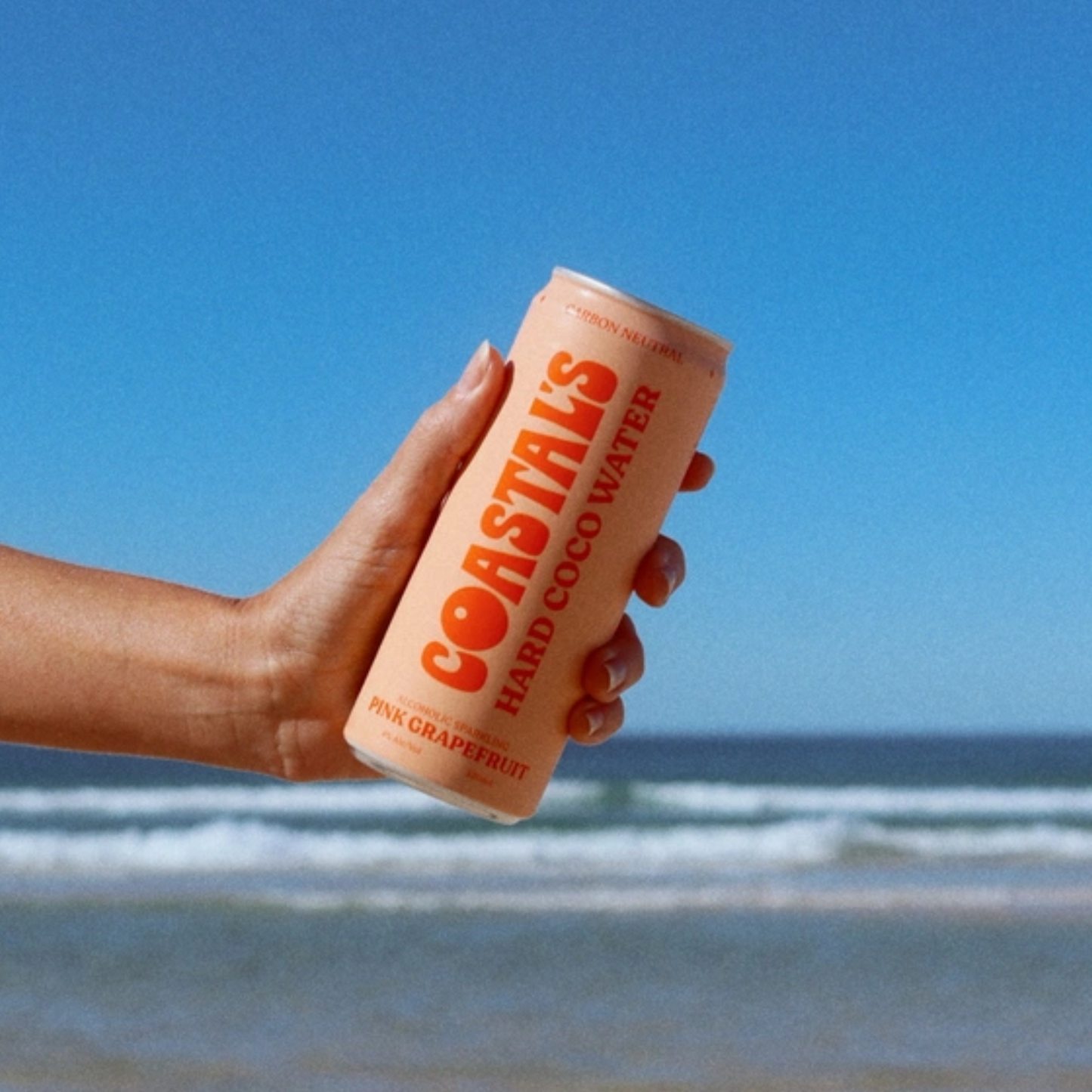 Hand holding a can of Coastals Hard Coconut water against a beach background