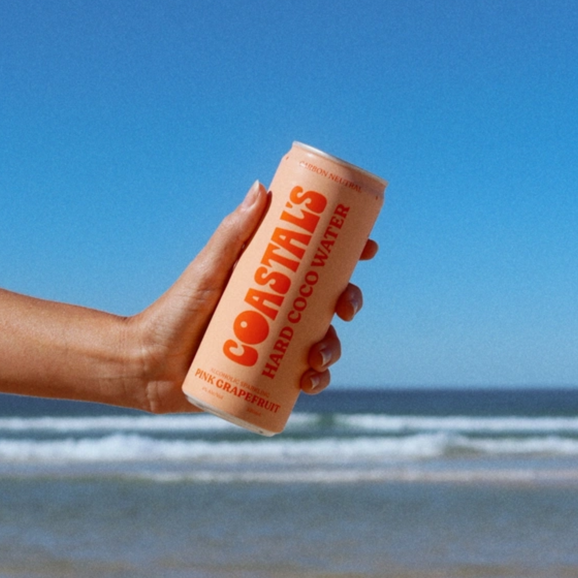 Hand holding a can of Coastals Hard Coconut water against a beach background